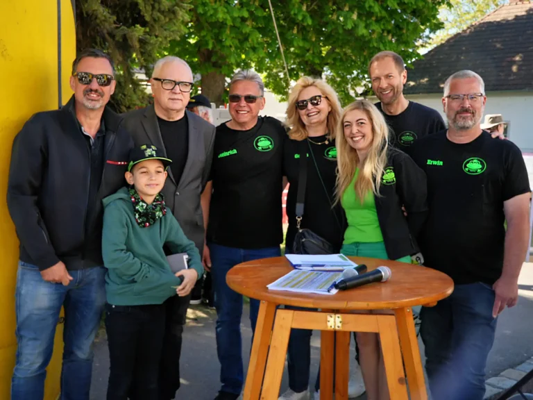 Alt-Text: Danke für diesen besonderen Tag Gruppenfoto eines Vereins- oder Organisationsteams bei einer Outdoor-Veranstaltung. Mehrere Erwachsene und ein Kind stehen lächelnd um einen Stehtisch mit Mikrofonen und Unterlagen. Einige tragen schwarze Shirts mit grünem Logo. Im Hintergrund sind Bäume und Gebäude bei sonnigem Wetter zu sehen.